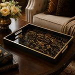 Gold baroque decorative tray on a dark wood coffee table, featuring intricate gold scrollwork on a black glass base with electroplated gold handles, styled with cream roses and antique books.