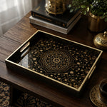 Islamic geometric decorative tray with a gold mandala and intricate arabesque floral vines on a black glass base, framed by a black tray with gold electroplated rim and handles, styled on a dark wood table with a Quran, brass lantern, and coffee cup.