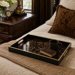 Baroque glass serving tray with gold scrollwork styled on a cream blanket, holding a black coffee mug and a vintage leather book, beside a dark wood nightstand and brown velvet pillow.