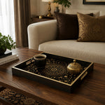 Gold and black Islamic arabesque tray on a dark wood coffee table, holding a black coffee mug and a small ornate brass box, with a cream sofa, brown velvet pillow, and patterned gold cushion in the background.
