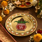 Gold Diwali charger plate with red kalash sacred pot, mango leaves, coconut, swastik symbol and scalloped red-green border for puja table.