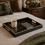 Gold and black decorative tray displayed on a cream linen bed, holding a white and gold coffee mug and a leather-bound book, with a green embroidered velvet pillow in the background.