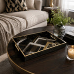 Geometric Art Deco decorative tray on a dark wood coffee table, with a lit candle, green foliage in a ribbed glass vase, and Interior Design books beside a cream sofa with a black and white diamond pillow.