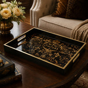 Gold baroque decorative tray on a dark wood coffee table, featuring intricate gold scrollwork on a black glass base with electroplated gold handles, styled with cream roses and antique books.