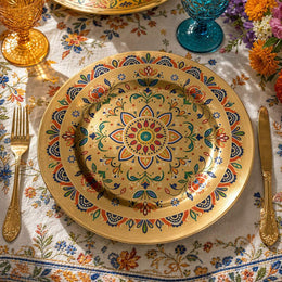 Gold electroplated Diwali charger plate with a multicolor rangoli mandala of lotus petals, paisley and small florals on a dining table.