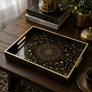 Islamic geometric decorative tray with a gold mandala and intricate arabesque floral vines on a black glass base, framed by a black tray with gold electroplated rim and handles, styled on a dark wood table with a Quran, brass lantern, and coffee cup.