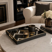 Black and gold Art Deco glass serving tray on a round white marble table, holding a black coffee mug and a small ribbed amber candle holder, with a cream armchair and white hydrangeas in the background.