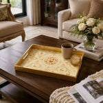 State of Qatar Glass Serving Tray – Gold Scrollwork Crest styled on a console table with a ceramic mug and books beside a brass lamp.
