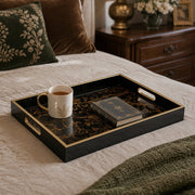 Gold and black decorative tray displayed on a cream linen bed, holding a white and gold coffee mug and a leather-bound book, with a green embroidered velvet pillow in the background.
