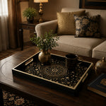 Islamic geometric mandala tray with gold filigree on a dark wood coffee table, displaying a small brass vase with greenery and a black and gold coffee cup, beside a cream sofa with gold and blue damask pillows.