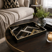 Geometric Art Deco decorative tray on a dark wood coffee table, with a lit candle, green foliage in a ribbed glass vase, and Interior Design books beside a cream sofa with a black and white diamond pillow.